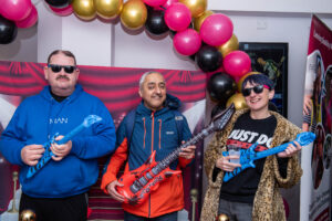2 men and a woman posing and looking cool with inflatable guitars and wearing sunglasses, under a balloon arch