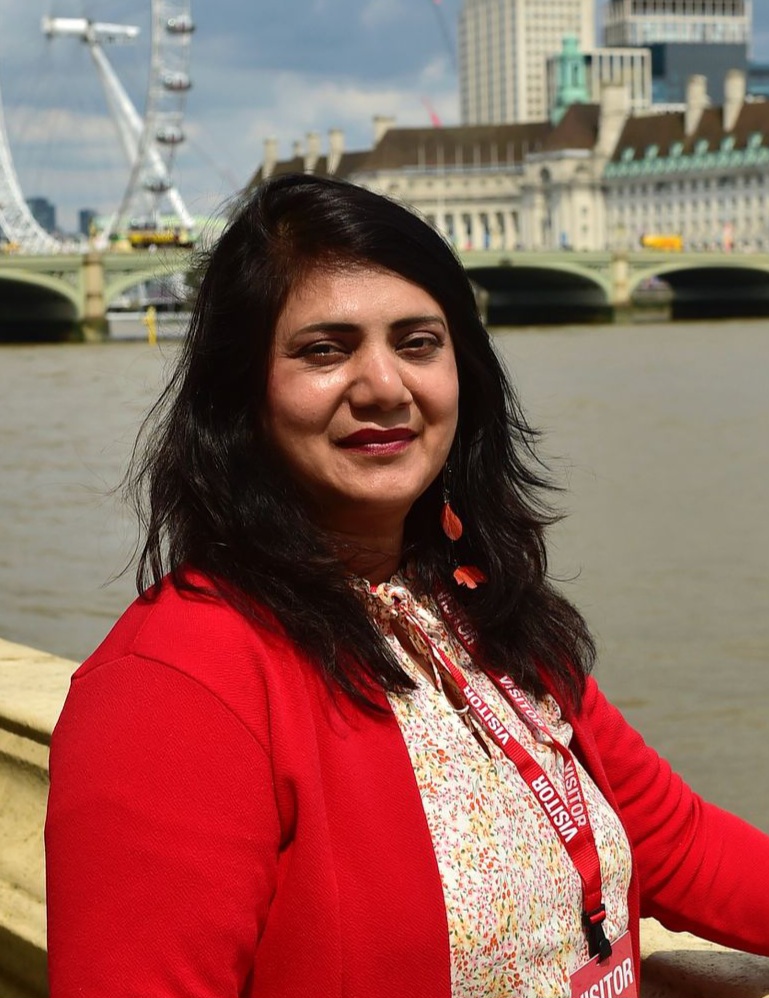 Julie has long black hair and is wearing a smart red cardigan and blouse. She is standing next to the Thames in Westminster with the London Eye behind her