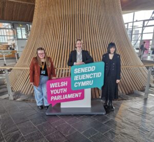 3 Welsh Youth Parliament Members including Georgia stood by the funnel in the Senedd holding a large cardboard Welsh Youth Parliament logo