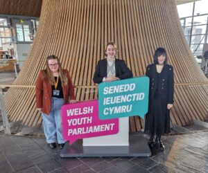 3 Welsh Youth Parliament Members including Georgia stood by the funnel in the Senedd holding a large cardboard Welsh Youth Parliament logo