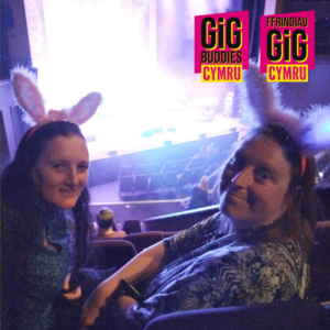 2 Gig Buddy friends at the theatre, they are turning around to the camera and smiling, with pink bunny ears on their heads, behind them is the glow from the stage lights